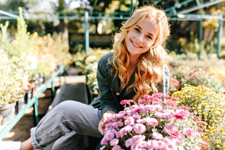Against background of bright flowering plants and green bushes that grow in garden beautiful blonde girl with green eyes smiles sweetly and basks in sun